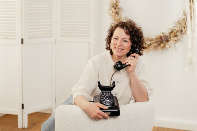Smiling woman sits on a sofa, holding a black rotary dial telephone to her ear.