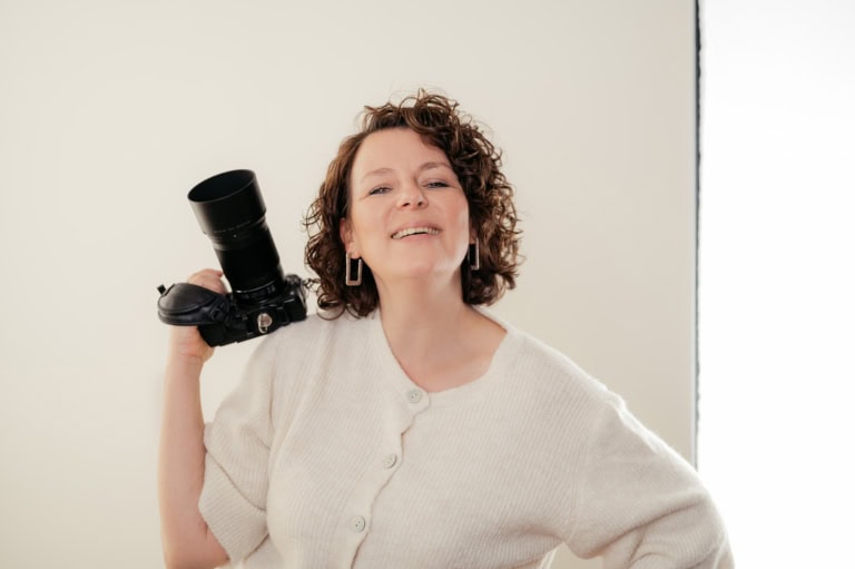 Smiling woman holding a DSLR with a large telephoto lens over her shoulder.