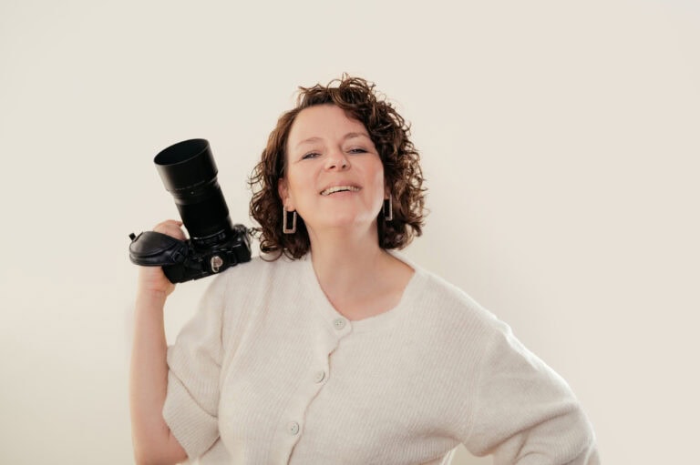 Smiling woman in a white sweater holds a large DSLR camera with a long lens over her shoulder against a pale background.
