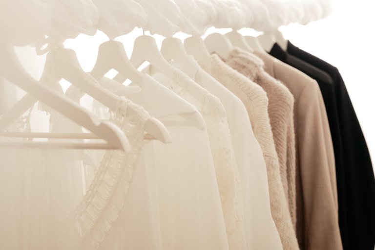 Rows of light-colored clothes on white hangers in a softly lit closet or wardrobe area