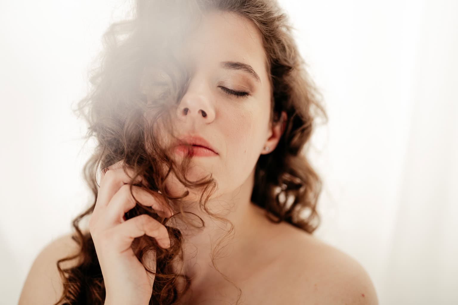 Close-up of a woman with eyes closed, touching her hair gently against a light, soft backdrop