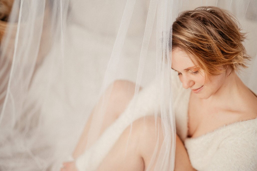 Close-up of a smiling woman with short hair, partially hidden behind a sheer white curtain, wearing a towel.