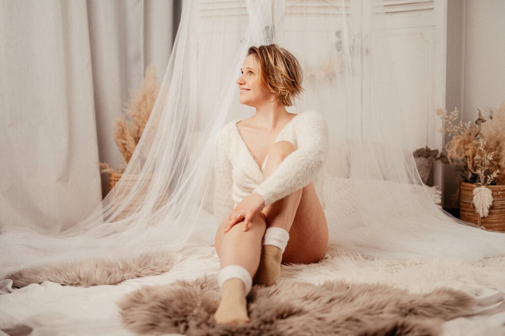 Woman with short blond hair sits on a bed in a fluffy white robe, a sheer veil draped over her, smiling softly.